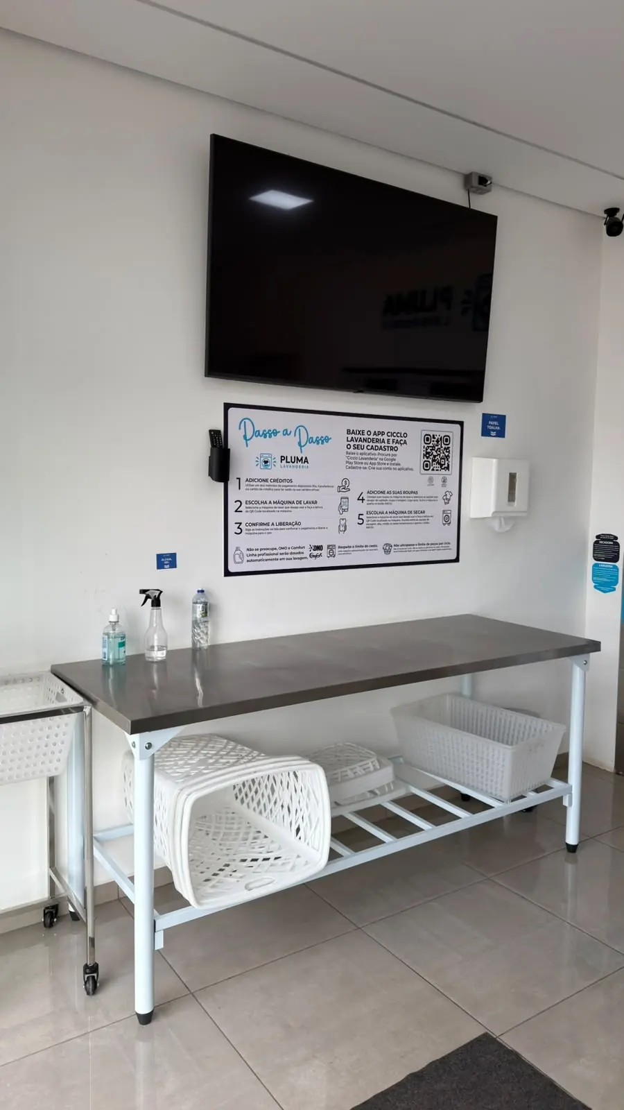Mesa de inox para dobrar roupa na Pluma Itaporanga, com cestos embaixo e o painel de passo a passo na parede.