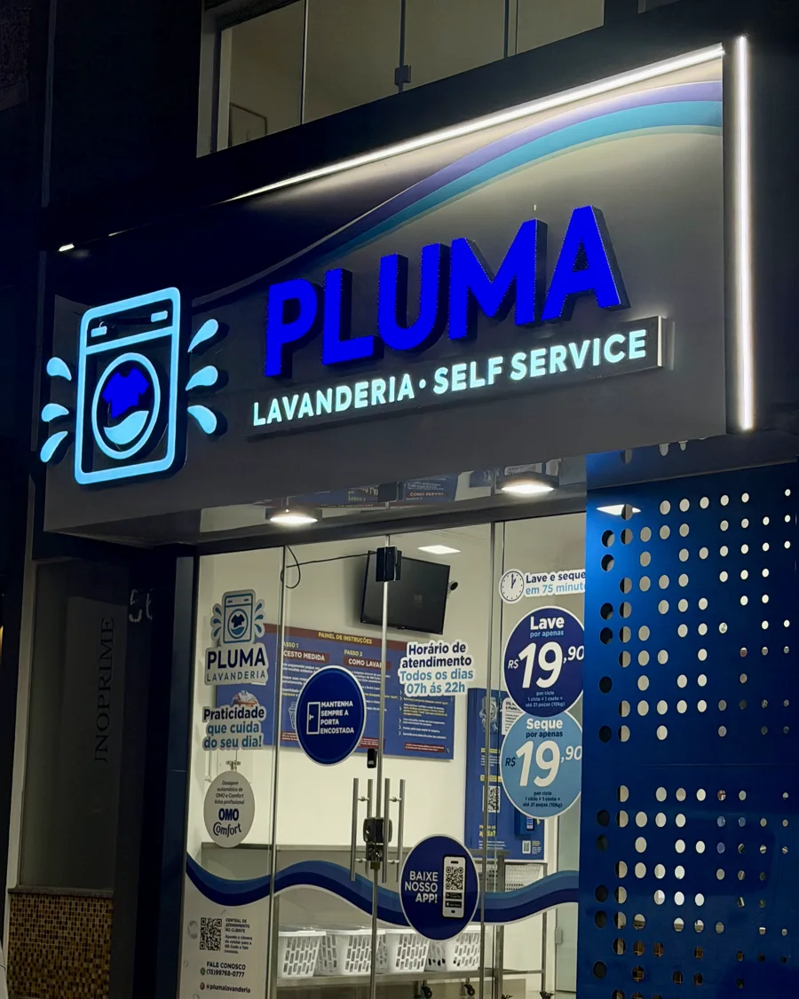 Fachada da Pluma Lavanderia em Itararé à noite: letreiro aceso em azul com a onda iluminada, e a vitrine mostrando os preços de R$ 19,90 para lavar e R$ 19,90 para secar e o horário das 7h às 22h.